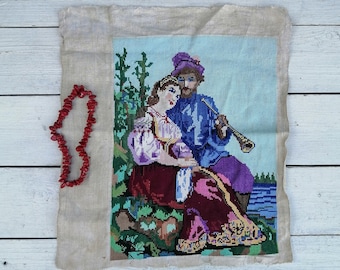 May include: A vintage needlepoint wall hanging featuring a couple sitting by a lake. The woman is wearing a purple dress and the man is wearing a blue shirt and a purple hat. The man is playing a flute.
