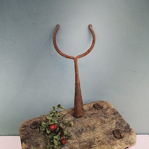 Op de afbeelding: Een roestige metalen haak met een puntig uiteinde, die op een rustieke houten kruk staat. De kruk heeft een verweerd oppervlak en is versierd met rode bessen en groene bladeren.