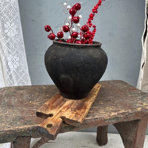 May include: A dark, textured, round-bellied cast iron pot filled with decorative stems featuring small, glossy red berries and white spherical accents. The pot sits on a weathered, rectangular wooden cutting board with a handle.