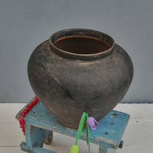 May include: A dark charcoal-colored ceramic pot with a wide, rounded body and a thick rim, resting on a small, distressed light blue wooden stool. Three small, colorful artificial tulip-shaped decorations in green, purple, and pink are attached.