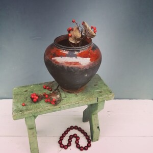 May include: A rustic brown clay pot with a dark glaze sits on a green wooden stool. The pot is filled with dried red berries and brown leaves. A strand of red beads lies on the white wooden floor.