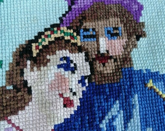 May include: A cross-stitch embroidery on light blue fabric depicting two figures. One figure wears a purple hat and a blue garment, while the other has brown hair with a colorful headband and a white patterned top. The stitching uses varied thread colors.