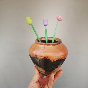 Op de afbeelding: Een hand houdt een klein, bruin aardewerken potje vast met een scheur die langs de zijkant loopt. Er zitten drie houten tulpen in de pot, een groene, een paarse en een roze.