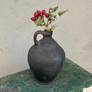 May include: A black ceramic pitcher with a wide mouth and a handle, filled with a sprig of red berries and green leaves, sitting on a green wooden table.