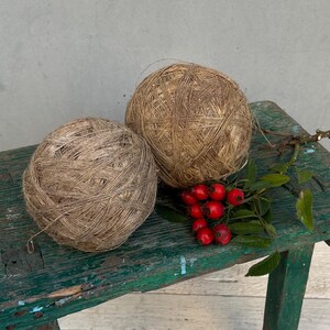 Op de afbeelding: Twee bollen natuurlijke vezelgaren, mogelijk jute of hennep, worden getoond op een groene houten bank. Het garen is lichtbruin van kleur en heeft een ruwe textuur. Een takje met rode bessen en groene bladeren is geplaatst tussen de bollen garen.