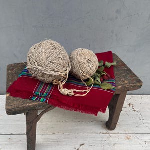 Op de afbeelding: Twee bollen beige garen liggen op een kleine, verweerde houten kruk. Een levendig rood textiel met kleurrijke strepen en franjes is over de kruk gedrapeerd. Gedroogde bessen en bladeren voegen een natuurlijk tintje toe.