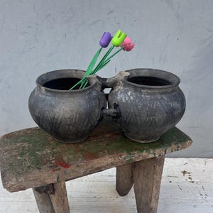 May include: Two black ceramic pots with a decorative design, connected together, sitting on a wooden stool with green and red paint.  Three wooden tulips in pink, purple, and green are in the pots.
