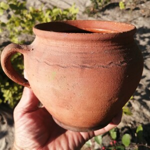 Pode incluir: Um pote de barro de terracota com uma alça. O pote é marrom e tem uma textura áspera. Está ligeiramente desgastado e tem alguns pequenos arranhões.