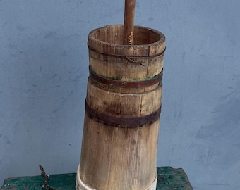 May include: A wooden butter churn with a wooden paddle, featuring a white band around the bottom and metal bands around the middle. The churn is sitting on a green wooden stool.
