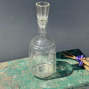 May include: A clear glass decanter with a textured surface and a stopper. The decanter is sitting on a green wooden surface.