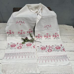 May include: White linen scarf with embroidered floral and bird motifs in red and gray. The scarf features a lace trim along the bottom edge. A sprig of dried roses rests on the scarf.