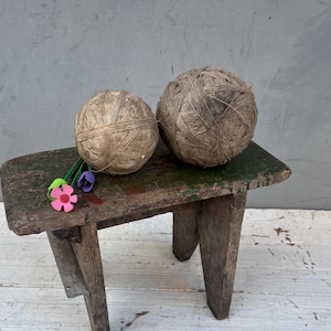 Può includere: Due gomitoli di spago, uno più grande dell'altro, sono appoggiati su uno sgabello di legno rustico. Lo sgabello è dipinto di verde e ha un aspetto usurato e invecchiato. Un piccolo fiore di feltro è sullo sgabello.