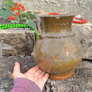 Può includere: Un vaso di terracotta marrone con un designo decorativo si trova su un ceppo d'albero. Il vaso è circondato da perline verdi e rosse.