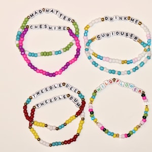 May include: Four beaded bracelets with white letter beads spelling "MAD HATTER", "CHESHIRE", "DRINK ME", "CURIOUSER", "TWEEDLE DEE", "TWEEDLE DUM", and "WILDFLOWER". The bracelets feature colorful beads in various hues, including pink, green, blue, yellow, and red.