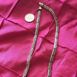 May include: A silver-toned necklace with a Greek key design, displayed on a pink fabric. The necklace is composed of rectangular links with a repeating geometric pattern. A US dime is next to the necklace for size reference.