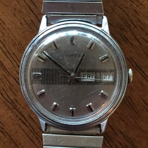 May include: A silver Timex wristwatch with a silver metal band. The watch face is silver with black hands and black Roman numeral hour markers. The watch has a day and date window that displays "FRI 27".