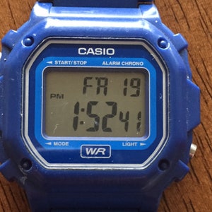 May include: A blue Casio digital wristwatch with a black face displaying the time, date, and day of the week. The watch has a silver band and a silver bezel. The watch face reads "FR 19 1:5241" and has the words "START/STOP", "ALARM CHRONO", "MODE", "LIGHT", and "WR" printed on it.