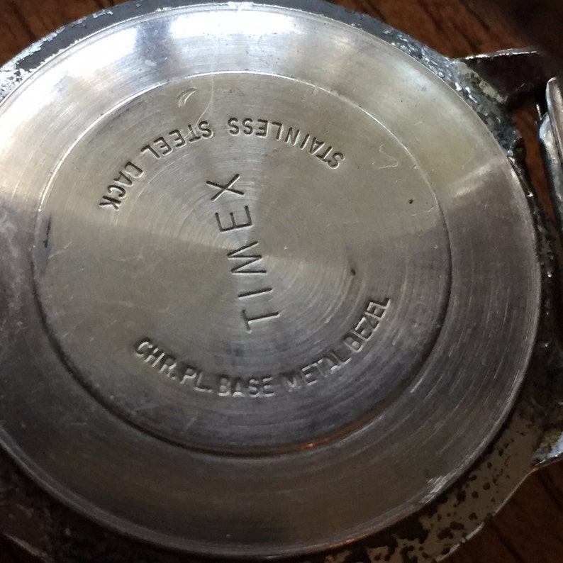 May include: The back of a silver Timex watch with the text "STAINLESS STEEL BACK", "TIMEX", "CHR. PL. BASE METAL BEZEL" engraved on it.