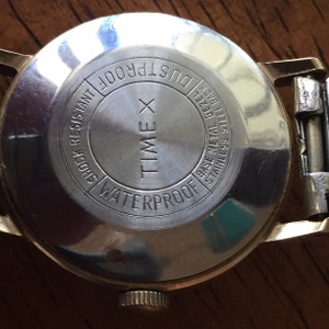 May include: The back of a gold Timex watch with the text "WATERPROOF", "TIME X", "DUSTPROOF", "SHOCK RESISTANT", and "BASE METAL BEZEL STAINLESS STEEL BACK" inscribed on the case.
