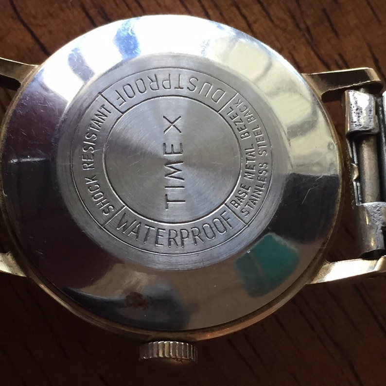 May include: The back of a gold Timex watch with the text "WATERPROOF", "TIME X", "DUSTPROOF", "SHOCK RESISTANT", and "BASE METAL BEZEL STAINLESS STEEL BACK" inscribed on the case.