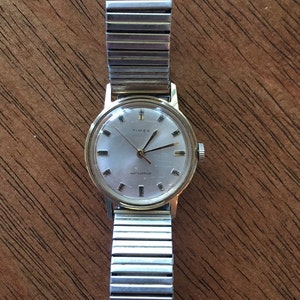 May include: A gold-tone Timex wristwatch with a silver metal band. The watch face is white with black markings and the words "Timex" and "Waterproof" printed on it.
