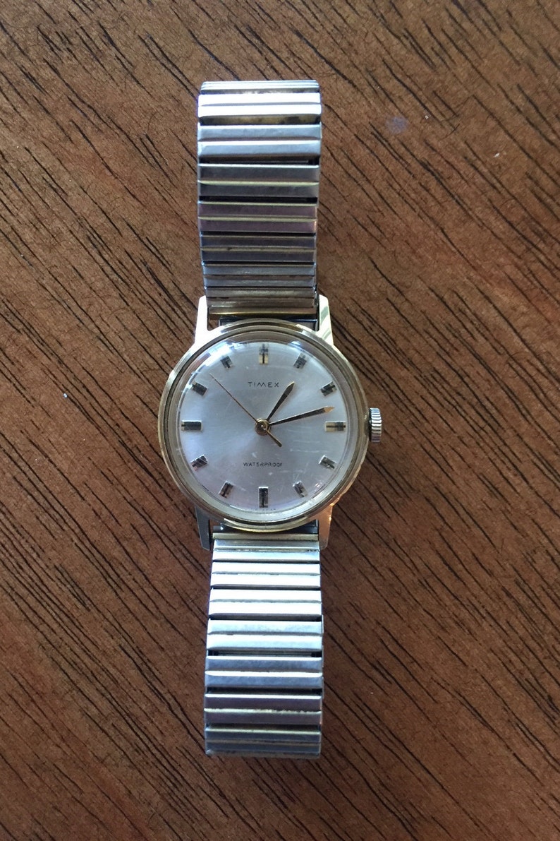 May include: A gold-tone Timex wristwatch with a silver metal band. The watch face is white with black markings and the words "Timex" and "Waterproof" printed on it.