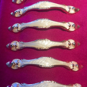 May include: Six ornate cabinet pulls with a white, pearlescent finish and gold accents. The pulls have a decorative, scrolled design and are arranged vertically on a pink background.