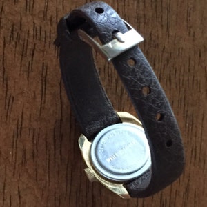 Pu&ograve; includere: Un orologio da polso dorato con un cinturino in pelle marrone. Sul retro dell'orologio &egrave; stampato il testo "WATER RESISTANT" e "STAINLESS STEEL".