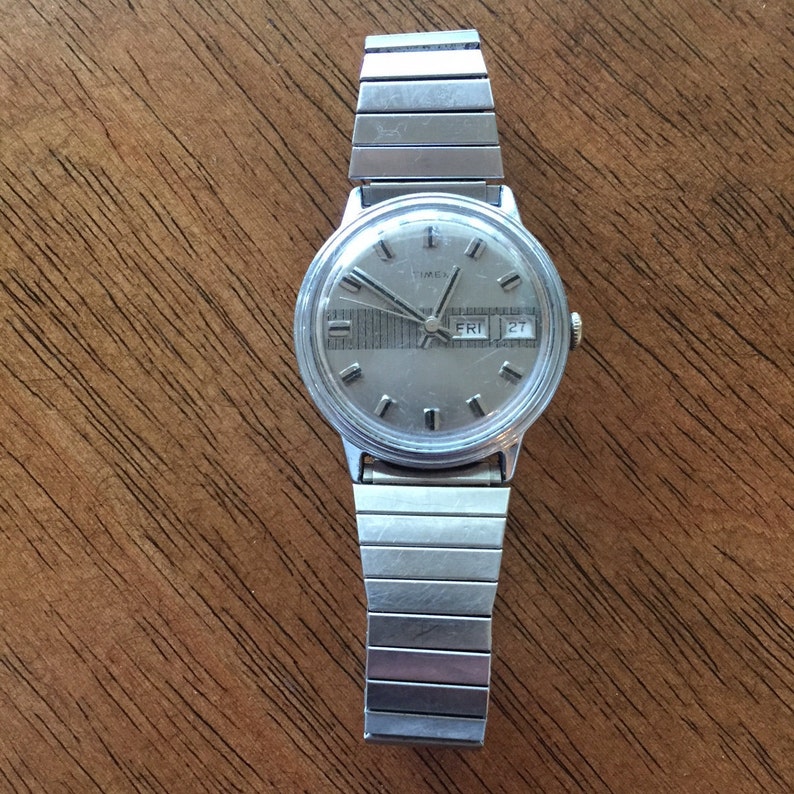 May include: A silver Timex wristwatch with a silver metal band. The watch face is silver with black markings and a day and date window. The day window shows "FRI" and the date window shows "27".