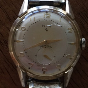 May include: A silver and gold men's wristwatch with a white face. The watch face has black Roman numeral hour markers and black minute markers. The watch has a gold second hand and a gold hour hand. The watch face also has the words "Elgin" and "Shockmaster" printed on it.