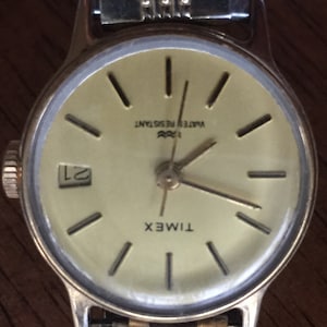 May include: A gold-toned Timex wristwatch with a white face and black hour and minute hands. The watch has a date window at the 3 o'clock position and the words "WATER RESISTANT" printed on the face. The watch is on a gold-toned metal band.