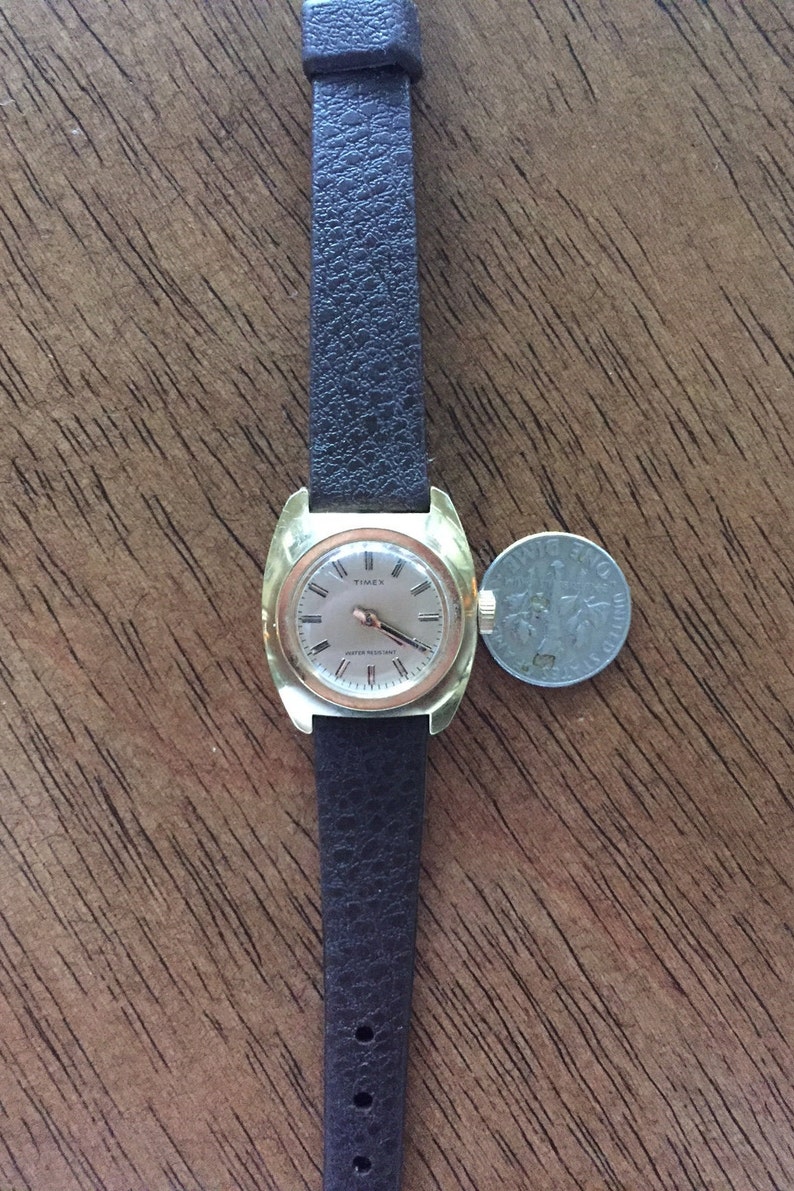 Pu&ograve; includere: Un orologio da polso Timex color oro con un cinturino in pelle marrone. L'orologio ha un quadrante quadrato con un quadrante bianco e lancette nere. L'orologio &egrave; resistente all'acqua.