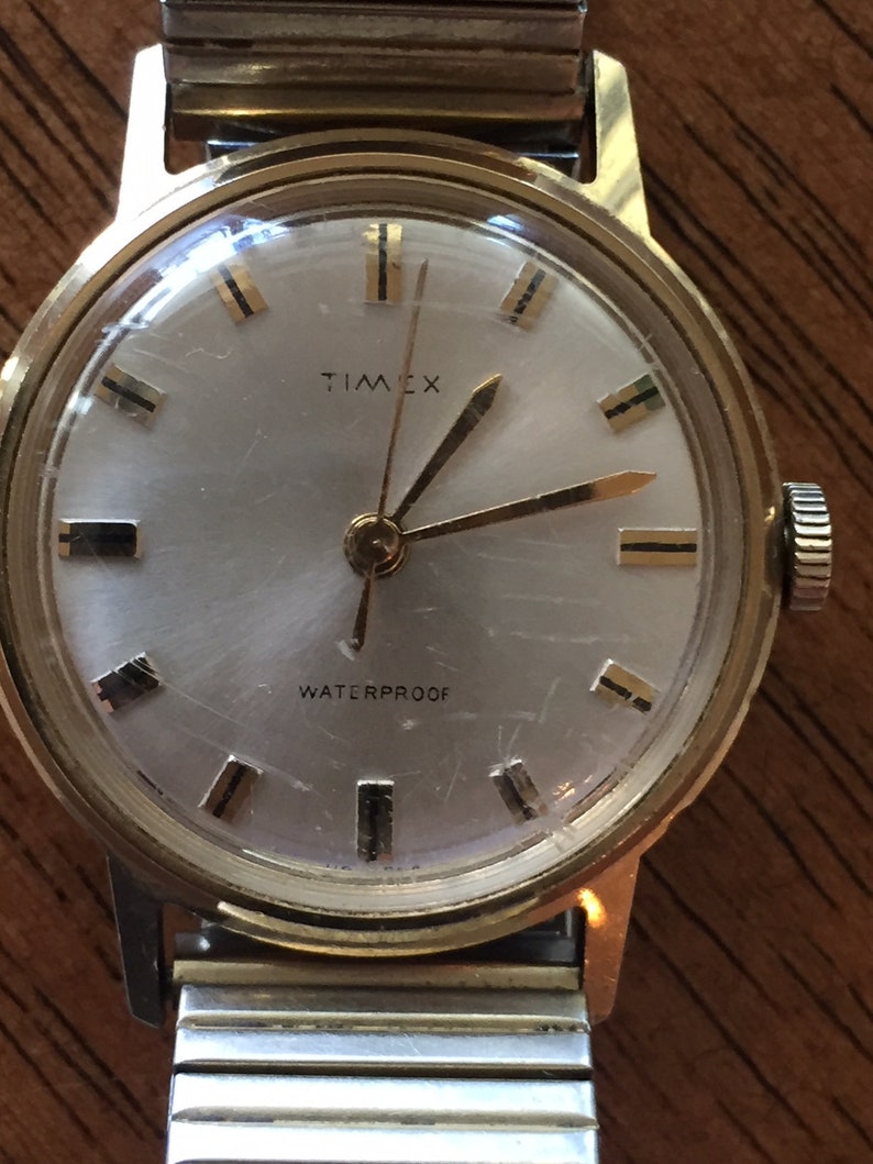 May include: A gold-toned Timex wristwatch with a white face and black hour markers. The watch face also says "WATERPROOF" and "TIMEX". The watch is on a silver and gold-toned metal band.