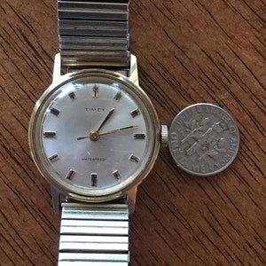 May include: A gold-toned Timex wristwatch with a white face and black hands. The watch has a silver metal band and says "Waterproof" on the face.
