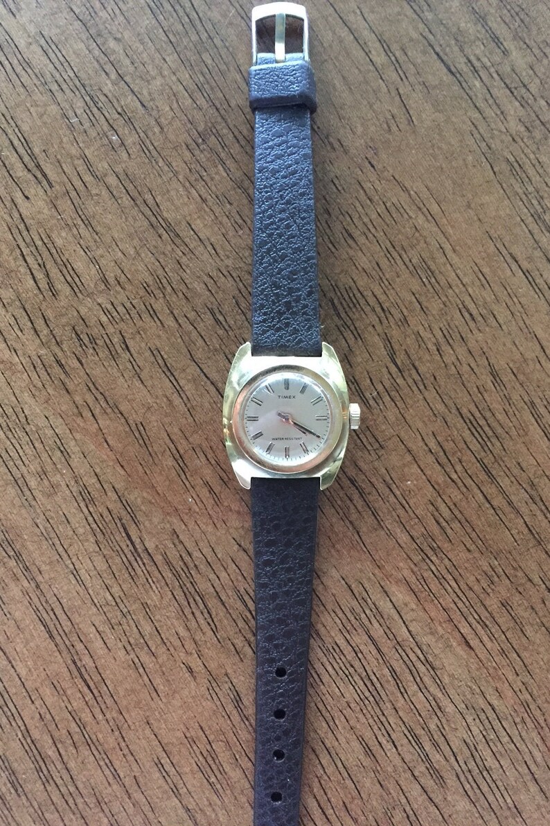Pu&ograve; includere: Un orologio Timex dorato con un quadrante quadrato e un cinturino in pelle nera. Il quadrante dell'orologio ha lancette e indici neri.