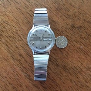 May include: A silver Timex wristwatch with a silver metal band. The watch face is silver with black markings and a day and date window. The day window shows "FRI" and the date window shows "27".
