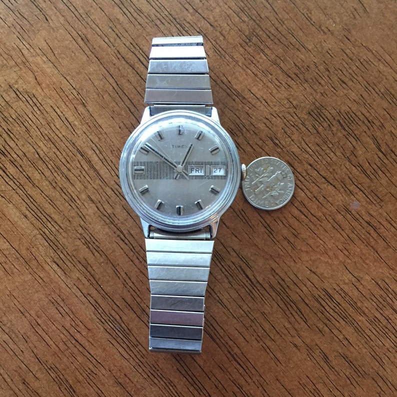 May include: A silver Timex wristwatch with a silver metal band. The watch face is silver with black markings and a day and date window. The day window shows "FRI" and the date window shows "27".