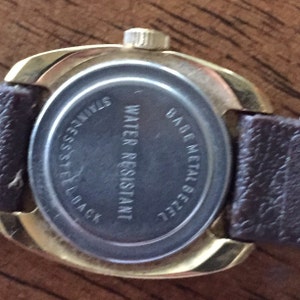 Pu&ograve; includere: Un orologio dorato con un cinturino in pelle marrone. Sul retro dell'orologio &egrave; stampato il testo "Water Resistant" e "Base Metal Back".