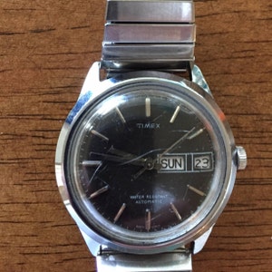 May include: A silver Timex watch with a black face, silver hands, and a date window that reads "SUN 23". The watch is water resistant and automatic.