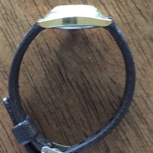Pu&ograve; includere: Un orologio da polso dorato con un cinturino in pelle marrone. L'orologio ha un quadrante quadrato e un design semplice.