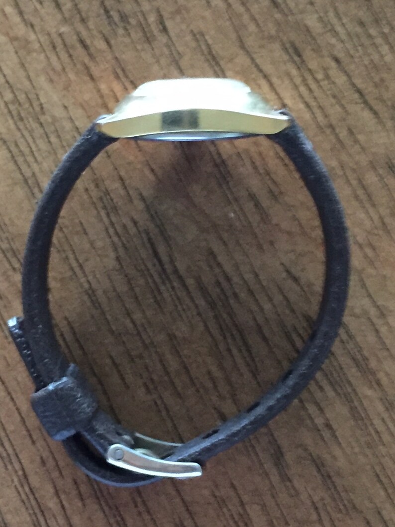 Pu&ograve; includere: Un orologio da polso dorato con un cinturino in pelle marrone. L'orologio ha un quadrante quadrato e un design semplice.