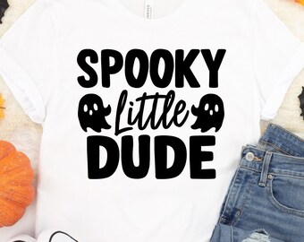 May include: A white short-sleeved t-shirt featuring the black text SPOOKY little DUDE with two ghost illustrations. The shirt is displayed alongside an orange pumpkin, blue denim jeans, orange canvas sneakers, and a plastic human skull.