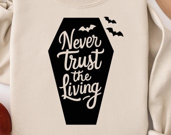 May include: A beige sweatshirt with the phrase "Never Trust the Living" in white script inside a black coffin shape. Three black bats are above the coffin. A fall-themed hat and pumpkin are in the background.