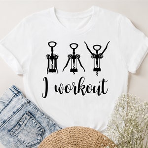 May include: White t-shirt with a black graphic of three corkscrews in different positions, with the text "I workout" below. The shirt is displayed on a light background with blue jeans and a woven hat.