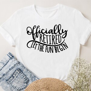 May include: White t-shirt with the text "Officially Retired, Let the Fun Begin" in black script. The shirt is folded, with a pair of blue jeans and a straw hat visible in the background. A sprig of white flowers is also present.
