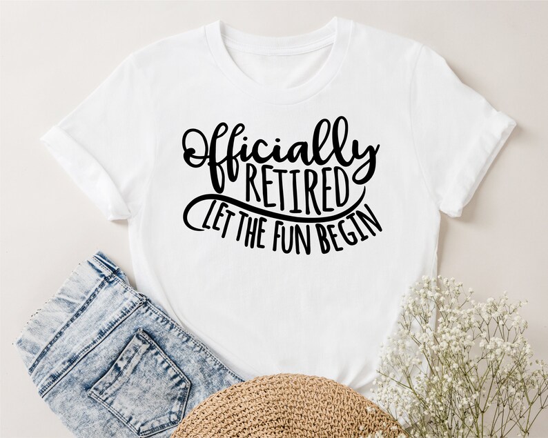 May include: White t-shirt with the text "Officially Retired, Let the Fun Begin" in black script. The shirt is folded, with a pair of blue jeans and a straw hat visible in the background. A sprig of white flowers is also present.