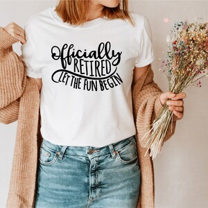 May include: White t-shirt with the text "Officially Retired Let The Fun Begin" in black script. The shirt is paired with blue jeans and a tan cardigan. A bouquet of colorful dried flowers is held in the hand.