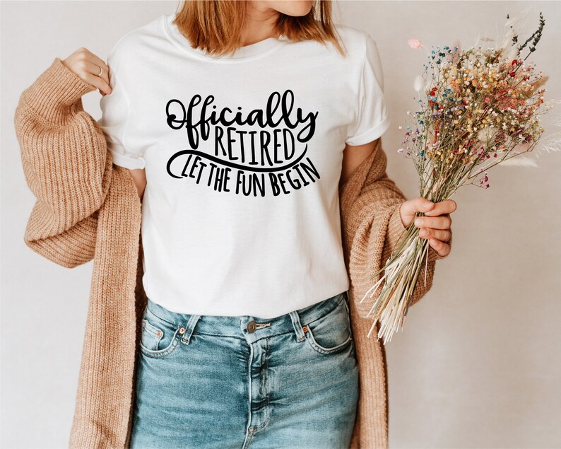 May include: White t-shirt with the text "Officially Retired Let The Fun Begin" in black script. The shirt is paired with blue jeans and a tan cardigan. A bouquet of colorful dried flowers is held in the hand.