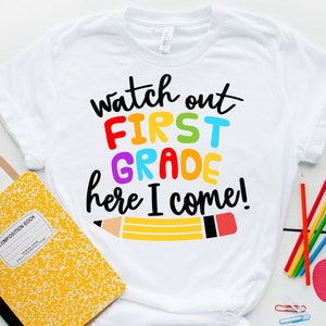May include: White t-shirt with the text "Watch out First Grade here I come!" in rainbow colors. A yellow pencil with a red eraser is drawn below the text.