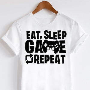 May include: White t-shirt with the text "EAT, SLEEP, GAME, REPEAT" in bold black letters. The word "GAME" features a black video game controller graphic. The shirt is hanging on a wooden hanger.